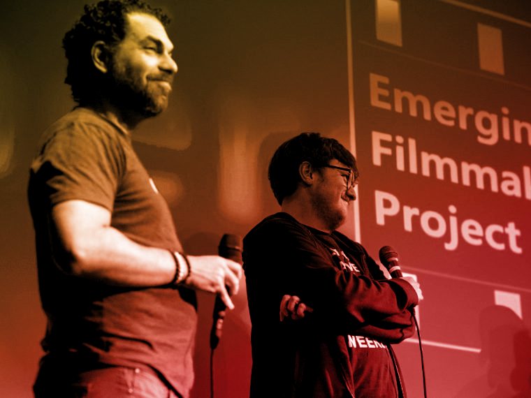 Dave Mercuri laughing on stage beside a collaborator, festival titles projected behind them